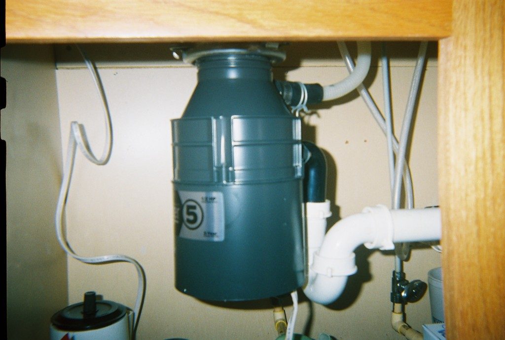 A gray garbage disposal unit installed under a kitchen sink with various white pipes and hoses connected to it, typically serviced by an emergency plumber in Atlanta.