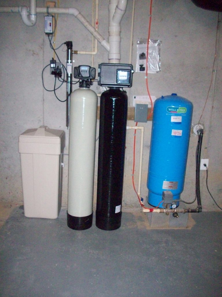 A basement water treatment system features two tanks (one beige, one black), a blue pressure tank, and associated control equipment and piping on a concrete floor. With our 24/7 plumbing service in Atlanta, you can ensure this setup remains effective and functional at all times.