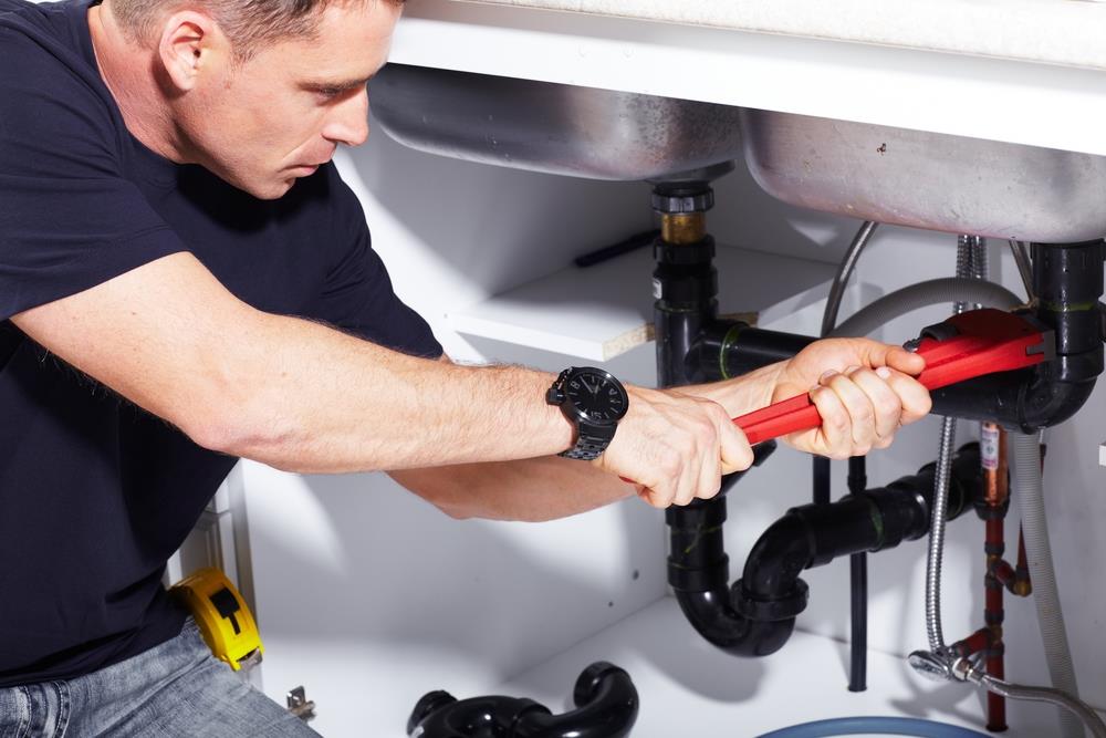 A person wearing a black shirt uses a red wrench to work on pipes under a sink, focusing on plumbing inspection and water heater repair.