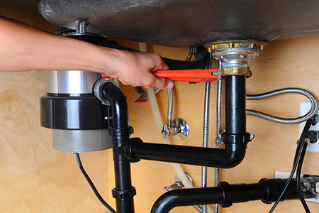 A person uses a red wrench to tighten a pipe joint under a kitchen sink with a garbage disposal unit installed, ensuring everything is secure during a plumbing inspection.