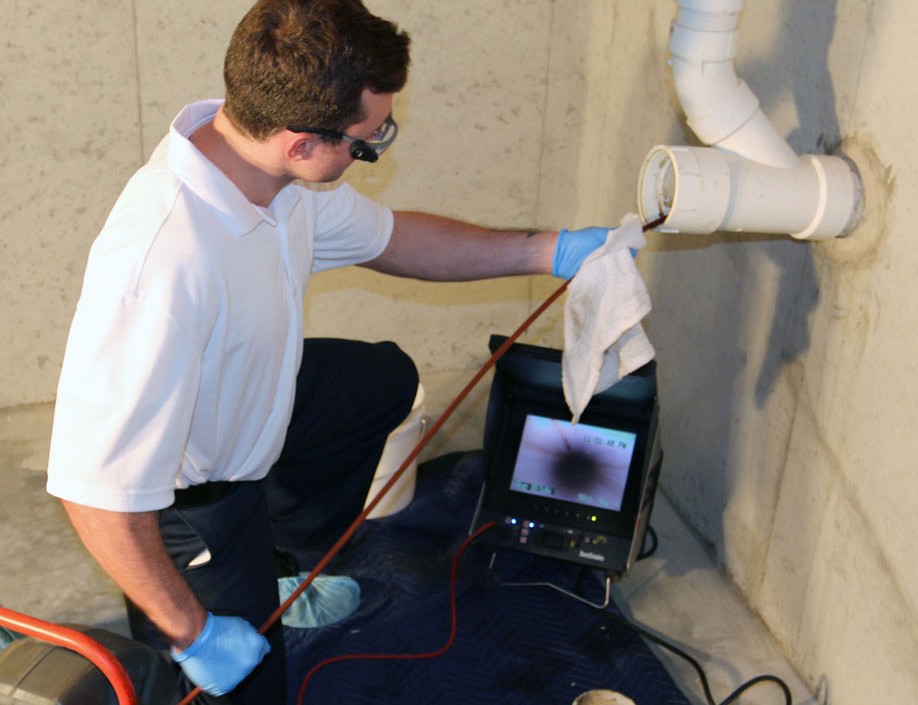 A person wearing gloves and safety glasses is inspecting a pipe for burst pipe repair using a camera system connected to a monitor. They are holding a cleaning cloth and operating equipment in a concrete basement.