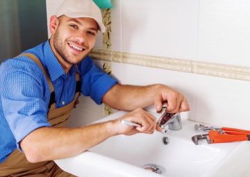 A person in work attire adjusts a faucet with a wrench in a bathroom setting, smiling at the camera, showcasing their skills in burst pipe repair.