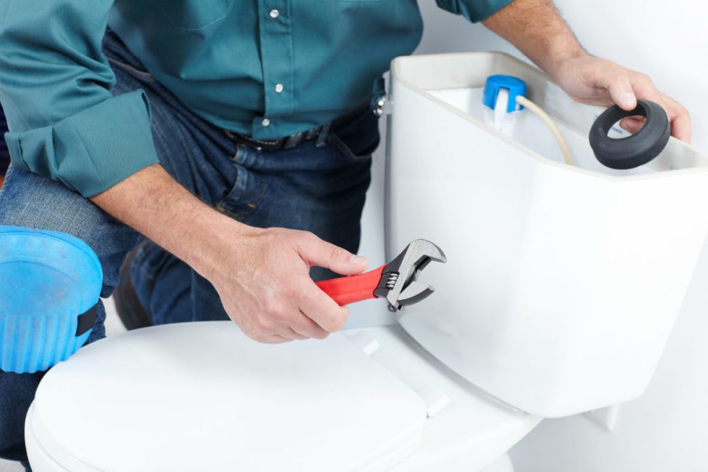 A person in a green shirt is using a wrench to repair a toilet tank for an emergency plumber in Atlanta.