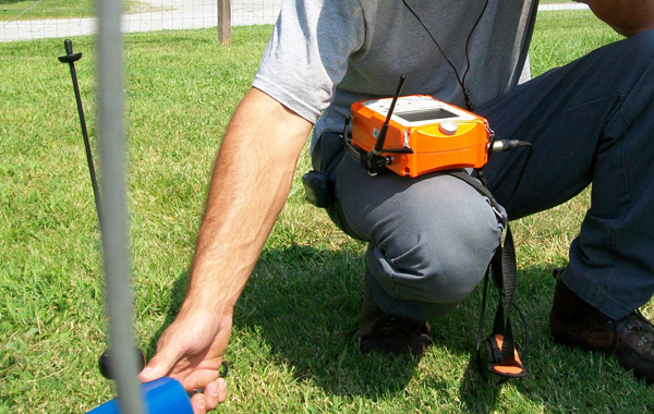 Person kneeling on grass, using a handheld underground utility locator with a bright orange device strapped to their waist, ensuring accurate placement during plumbing inspection.