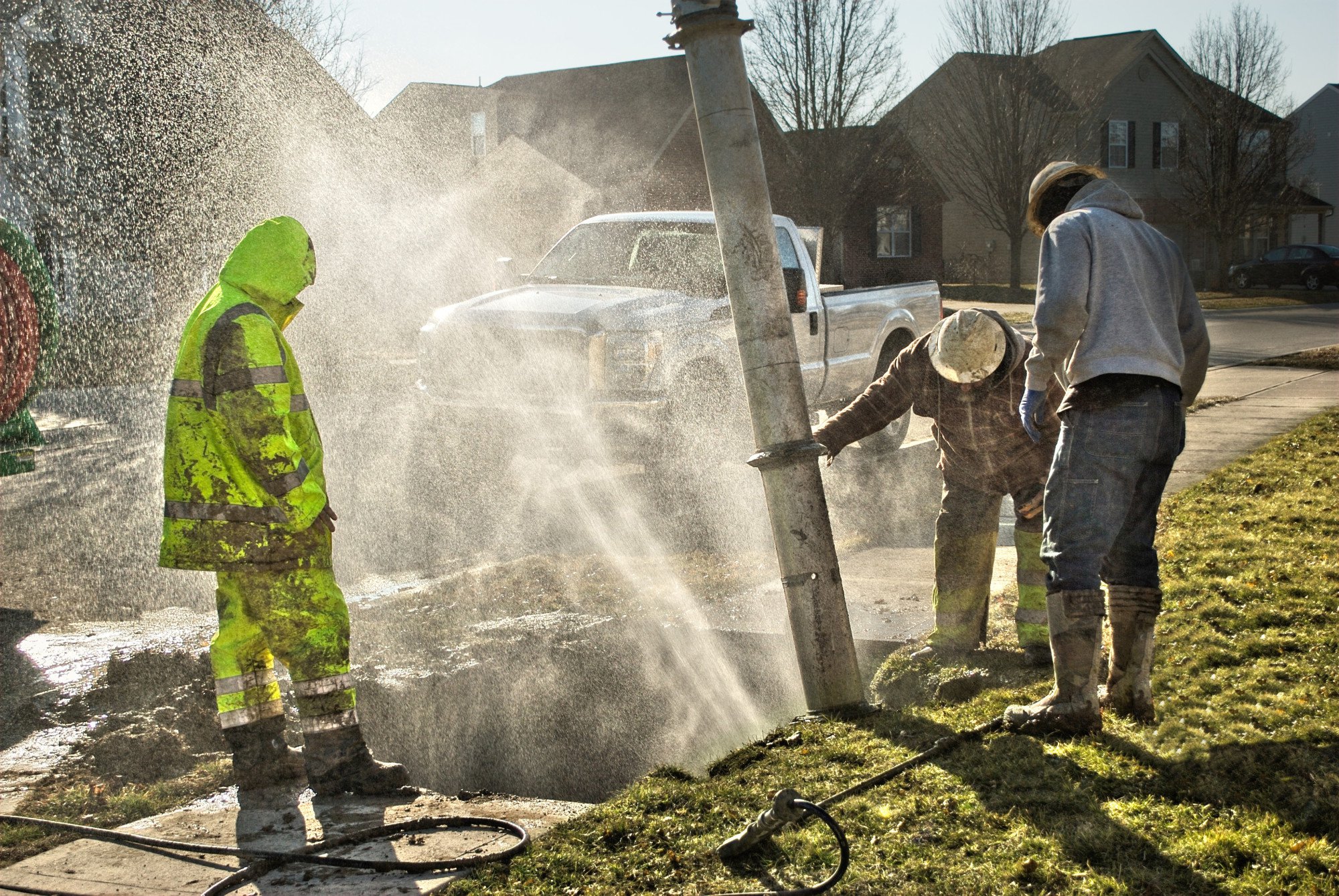 Three workers inspect a leaking fire hydrant on a residential street, with water spraying from the hydrant due to a burst pipe. A white truck is parked in the background.