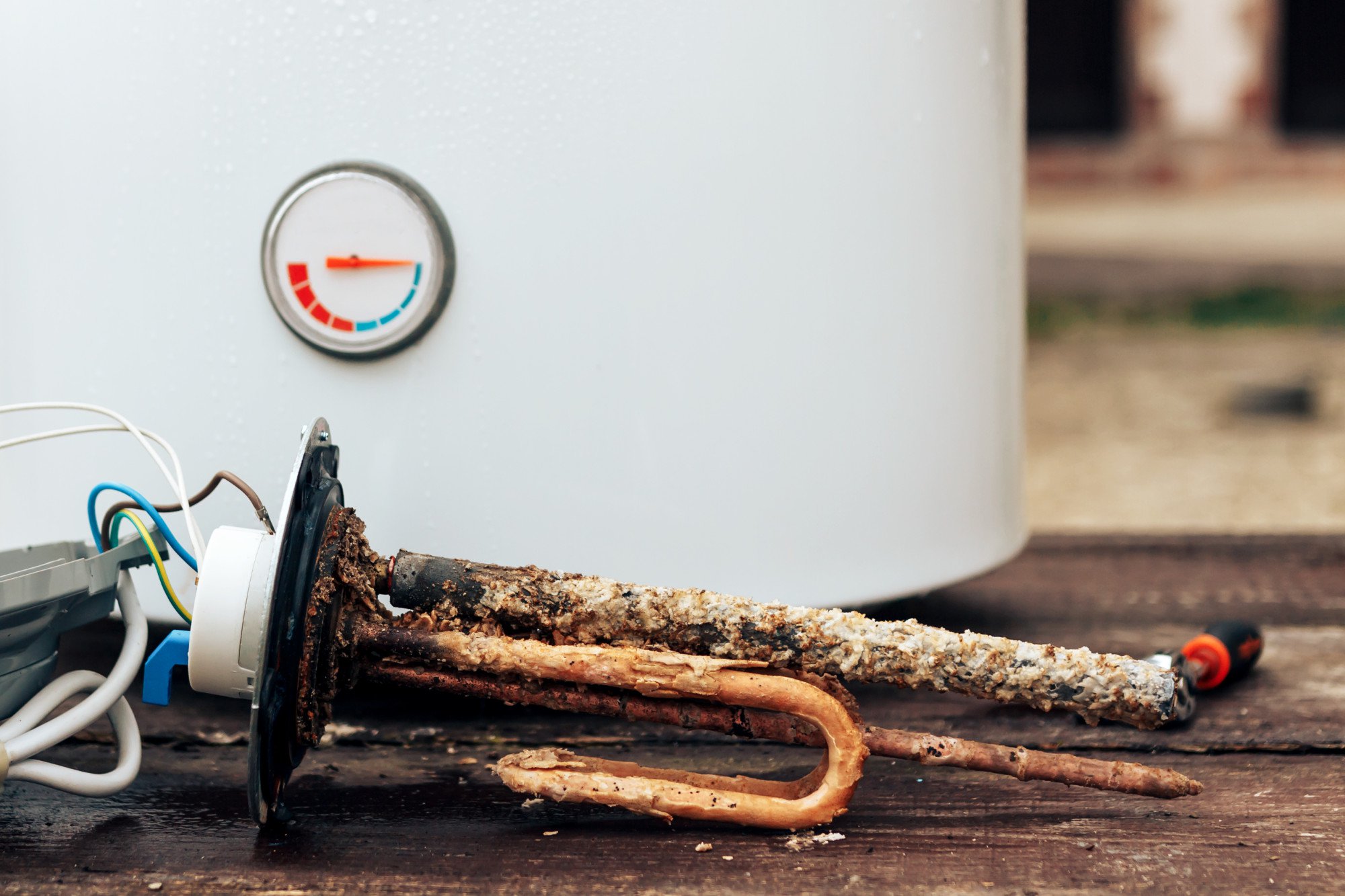 Close-up of a corroded water heater heating element lying on the ground in front of a white water heater with a temperature gauge, highlighting the need for water heater repair.