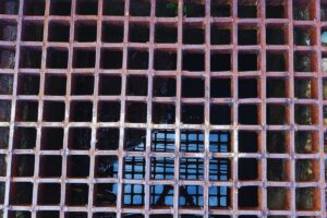 Rusty metal grate covering a square sewage drain with water visible beneath.